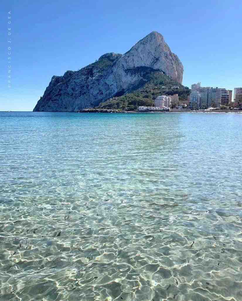 Clear turquoise waters exemplify the Mediterranean Sea, gently reflecting the sunlight. The prominent Peñón de Ifach rises dramatically in the background, surrounded by lush greenery and the coastal town below, creating a serene and picturesque natural setting.