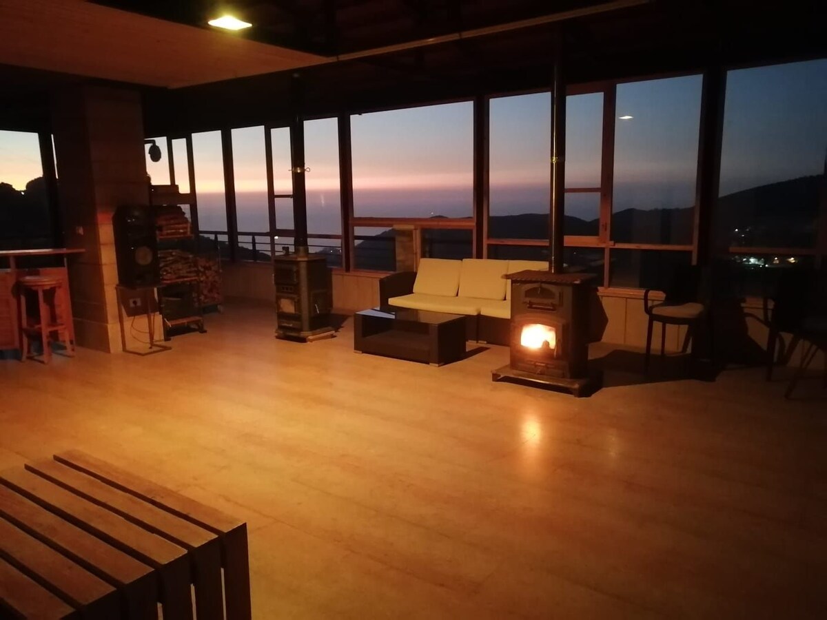 An open lounge area is showcased, featuring large windows that provide expansive views of the sea and mountains at sunset. A warm glow from a rustic stove creates a cozy corner with modern seating arrangements, enhancing the inviting environment.