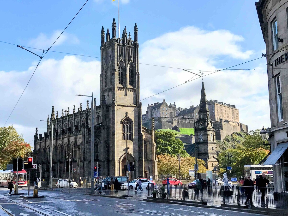 A view showcases St Mary’s Cathedral with its three prominent spires, framed by the historic architecture of the surrounding buildings. The iconic Edinburgh Castle looms in the background, set against a partly cloudy sky, while tram tracks run along the street in the foreground.