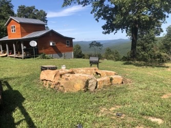 An outdoor fire pit constructed from natural stones is set in a grassy area. The fire pit is surrounded by a view of rolling hills and expansive trees, with a rustic cabin visible in the background against a clear blue sky.
