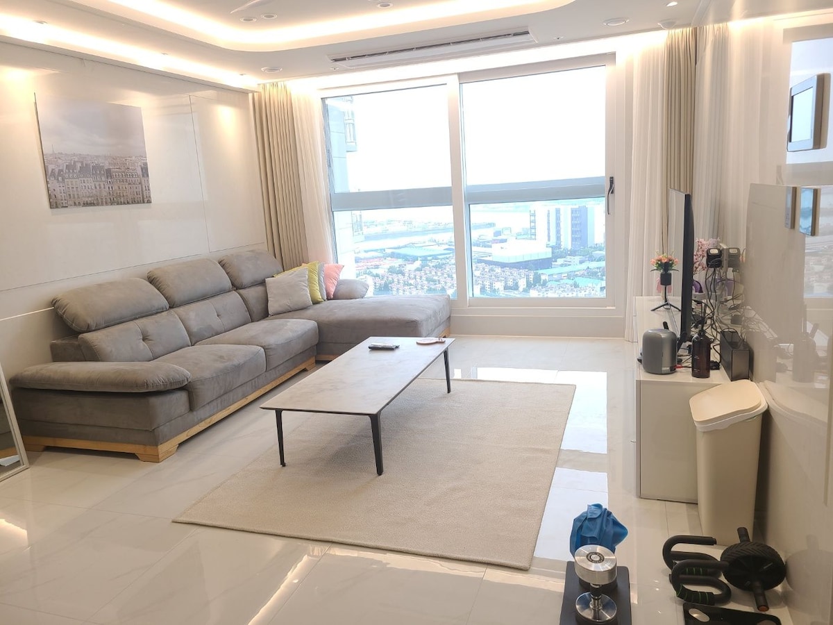 A spacious living room is highlighted by a large, L-shaped sofa and a low coffee table on a soft area rug. Natural light fills the space through a large window, offering views of the surrounding area. Modern furnishings and a sleek design are evident.