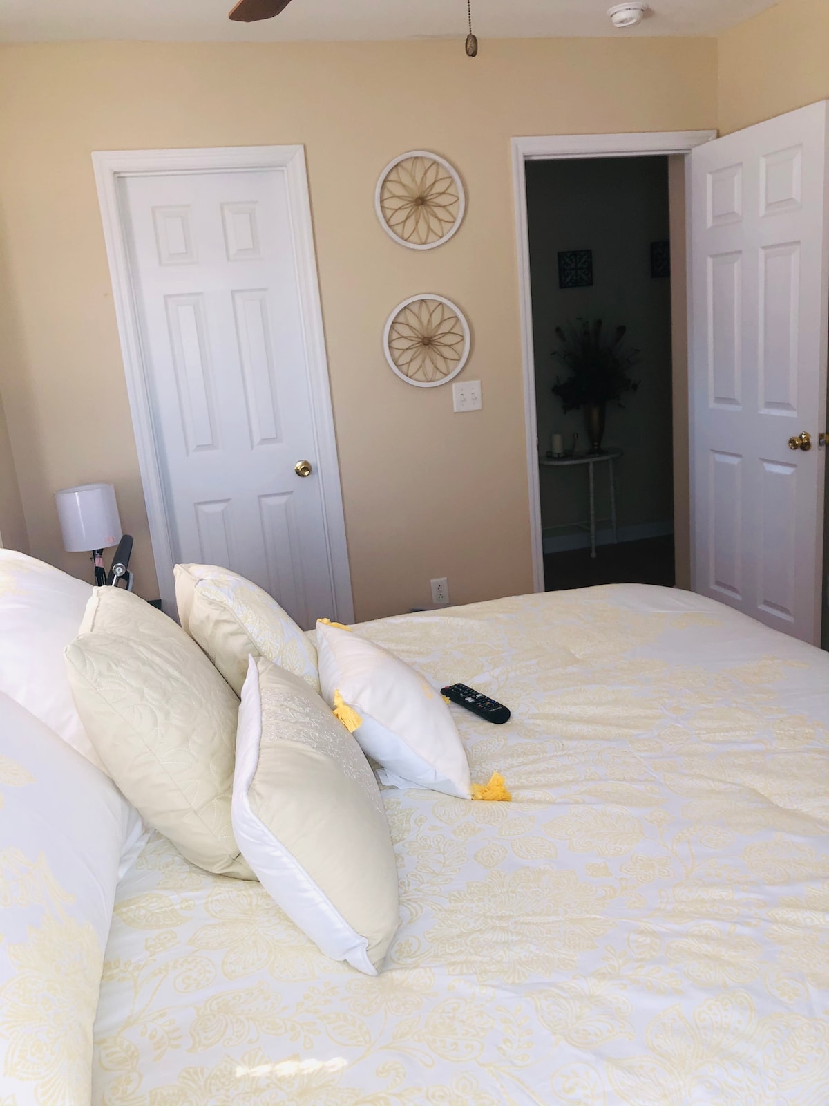 A cozy bedroom is adorned with light-colored bedding and decorative pillows. Two doors lead to other areas of the townhouse, while circular wall decor enhances the welcoming ambiance. A bedside lamp and remote control are placed within reach on the bed.