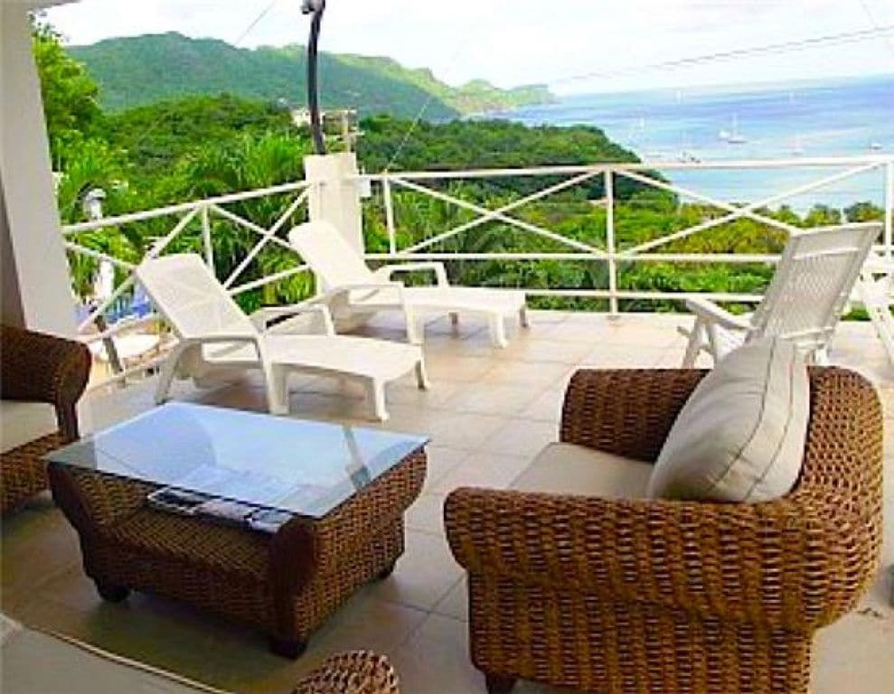 An outdoor lounge area features a glass-top table and comfortable wicker seating arranged for relaxation. Sun loungers are positioned nearby, overlooking lush green hills and a serene bay with sailboats, providing a tranquil setting for enjoying the views.