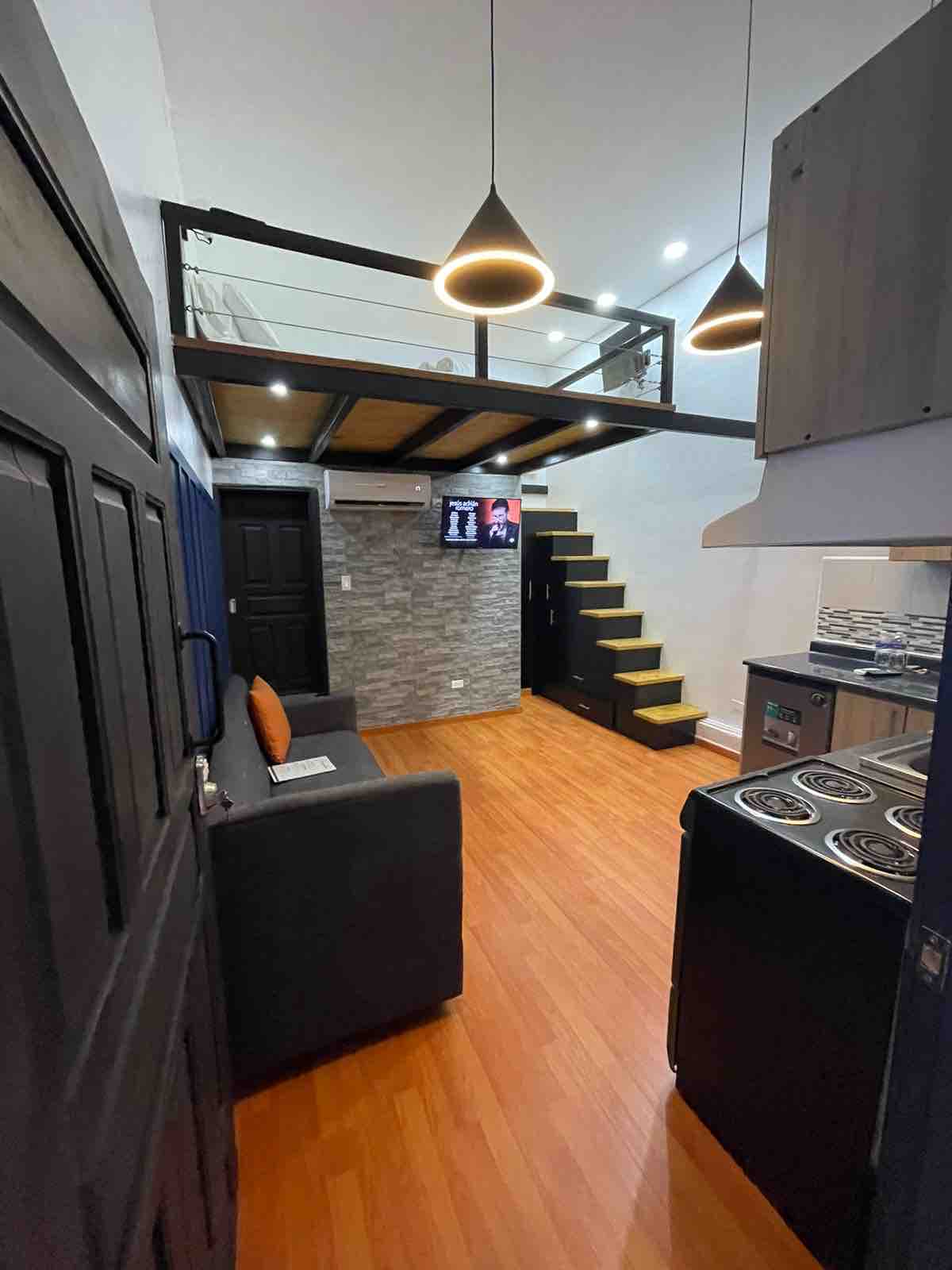 A contemporary living space is highlighted by warm wooden flooring and a sleek kitchen area. A staircase leads to an upper loft, while modern pendant lights hang above. A sofa and TV are positioned for relaxation, with a feature wall adding texture to the room.