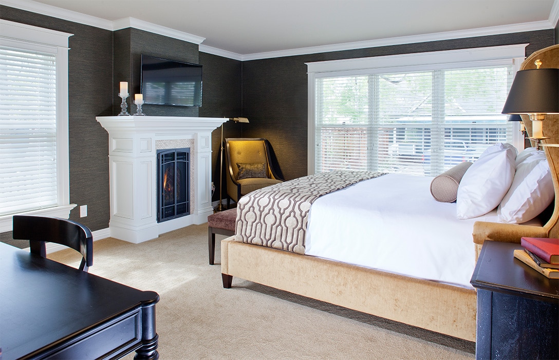 A spacious room features a kingsize bed with luxury linens and a cozy throw. A gas fireplace is positioned near a sitting area with a comfortable chair. Natural light streams in through large windows, illuminating warm-colored walls and neutral carpeting.