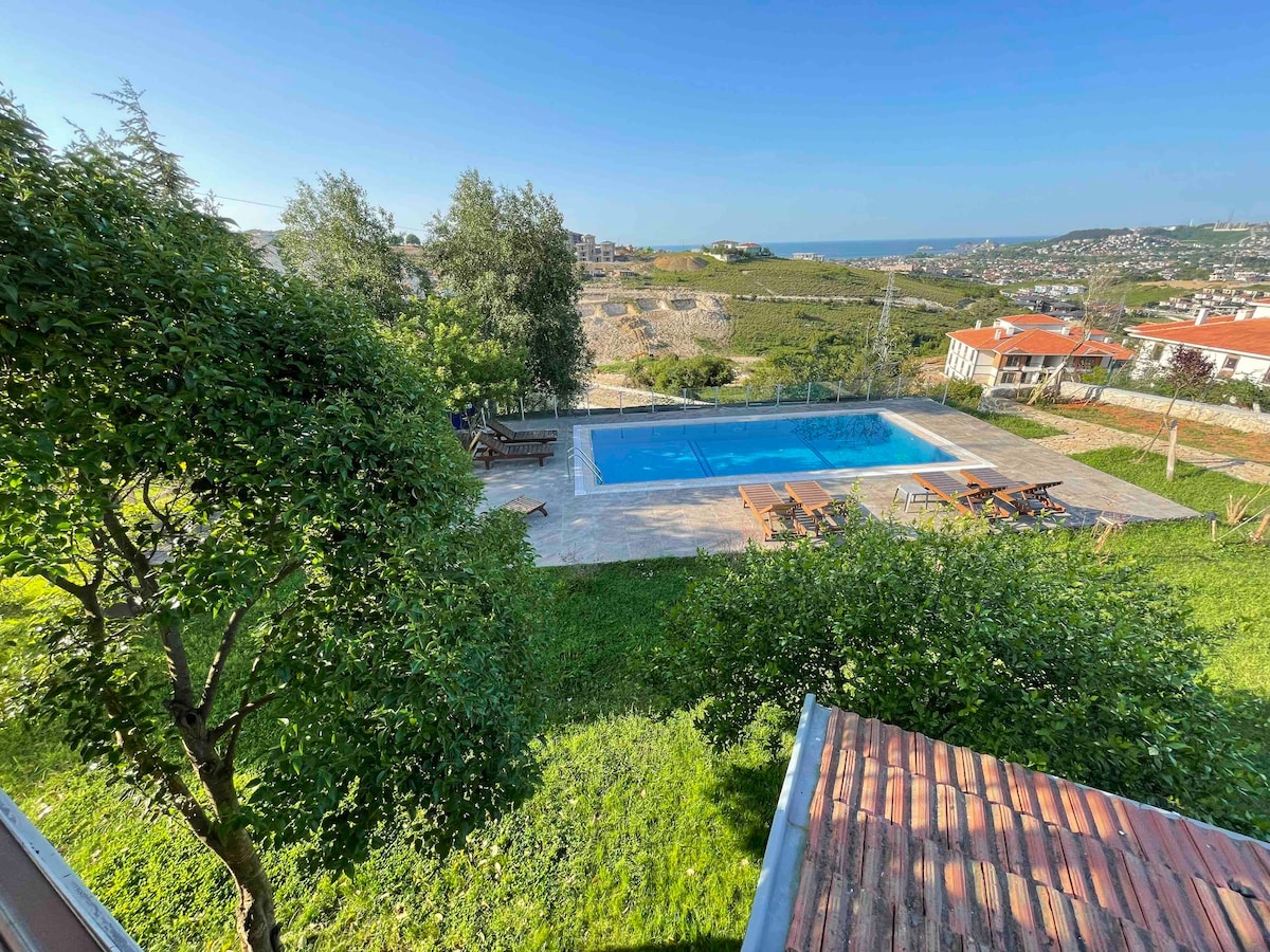 A serene outdoor area features a blue swimming pool surrounded by stone paving, set within lush green gardens. Wooden lounge chairs and a dining table are arranged nearby, offering a view of the expansive landscape beyond. Clear skies provide bright natural light.