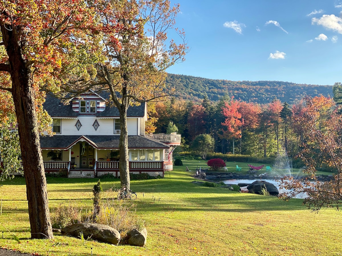 A charming home is set against a backdrop of vibrant autumn foliage and rolling hills. The exterior features a wraparound porch and is surrounded by a spacious lawn, while a serene lake is visible nearby, enhanced by natural landscaping and nearby seating areas.