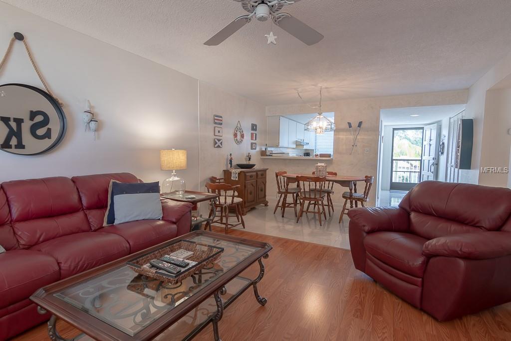 A spacious living area features a blend of leather seating and a coffee table, with light wood accents. A dining table for four is seen in the background, accompanied by chairs. Outdoor light filters in through a nearby entryway, enhancing the open atmosphere.