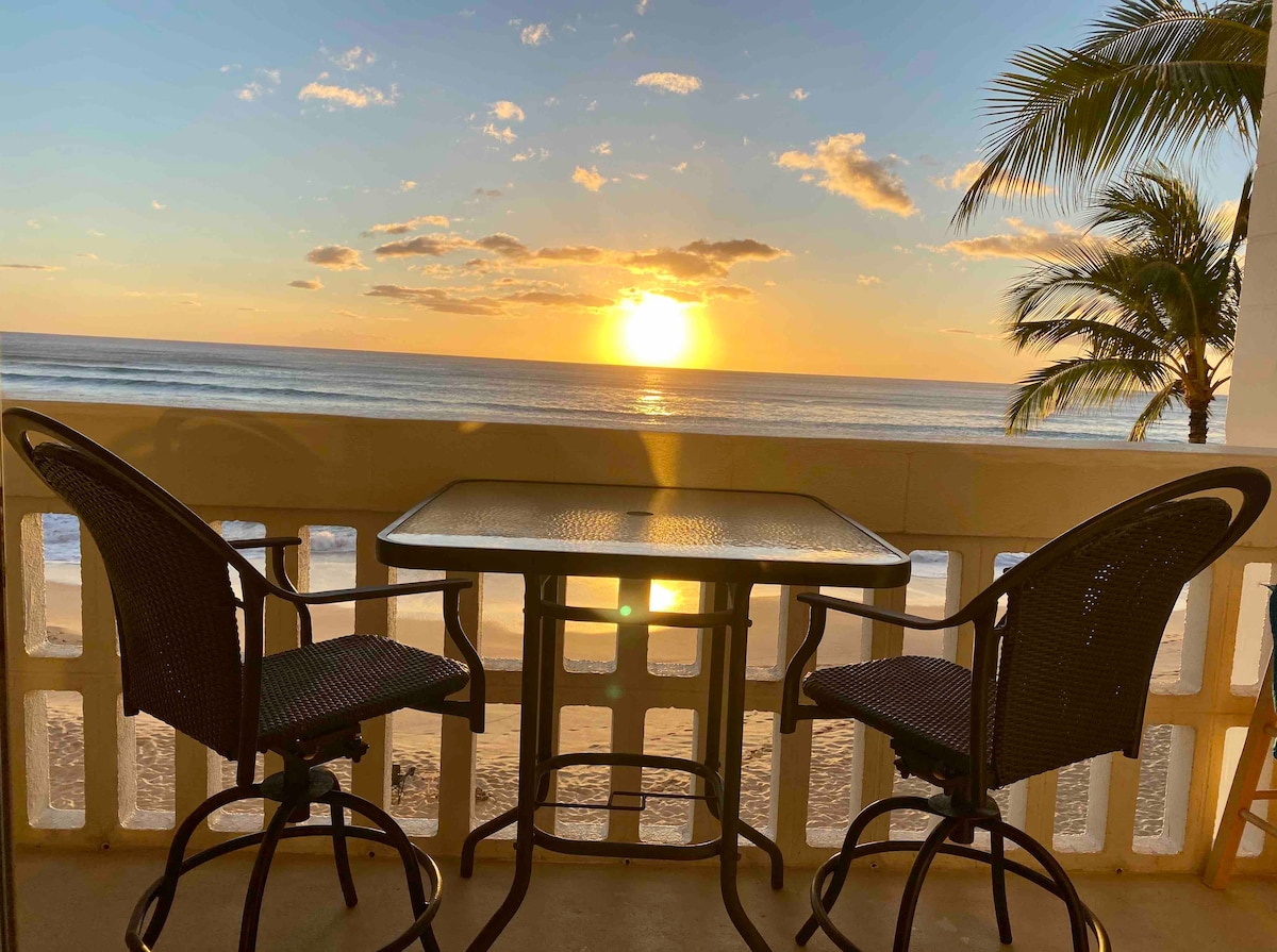 A private lanai is featured overlooking the beach, showcasing a table with two chairs. The sun sets on the horizon, casting warm colors across the sky and reflecting off the ocean's surface.
