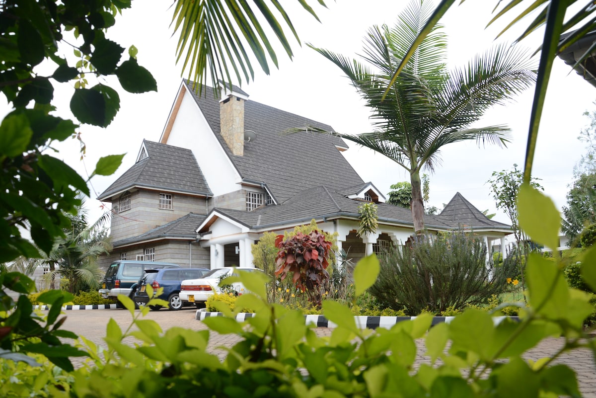 A spacious main house is framed by lush greenery, featuring a gabled roof and multiple architectural elements. Surrounding plants and trees enhance the outdoor aesthetic, while a paved driveway accommodates parked vehicles alongside the ample garden space.