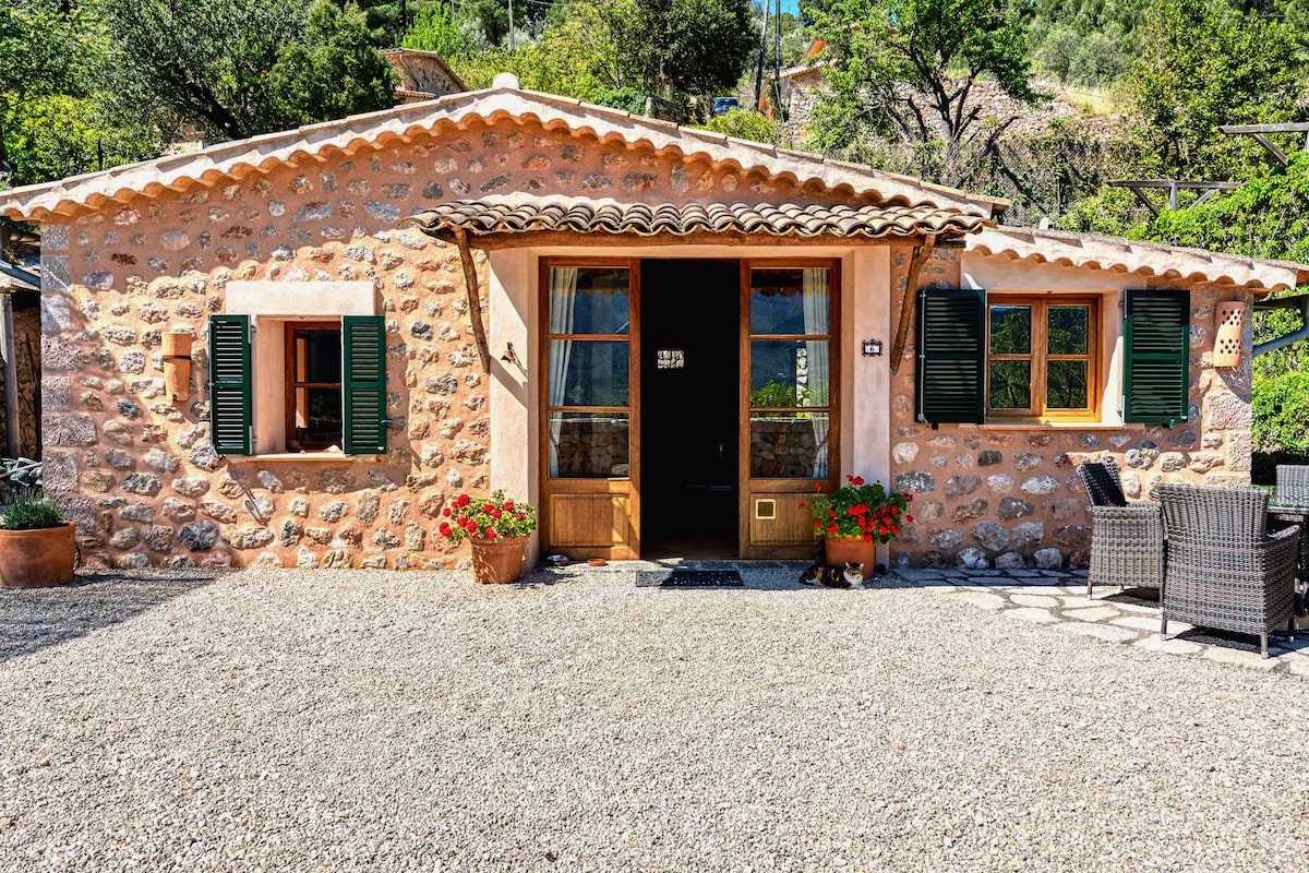 A charming stone cottage features a rustic exterior with brown shutters and a tiled roof. Potted flowers add color beside the gravel pathway leading to double doors. A seating area with wicker chairs is positioned on the front patio, surrounded by natural greenery.