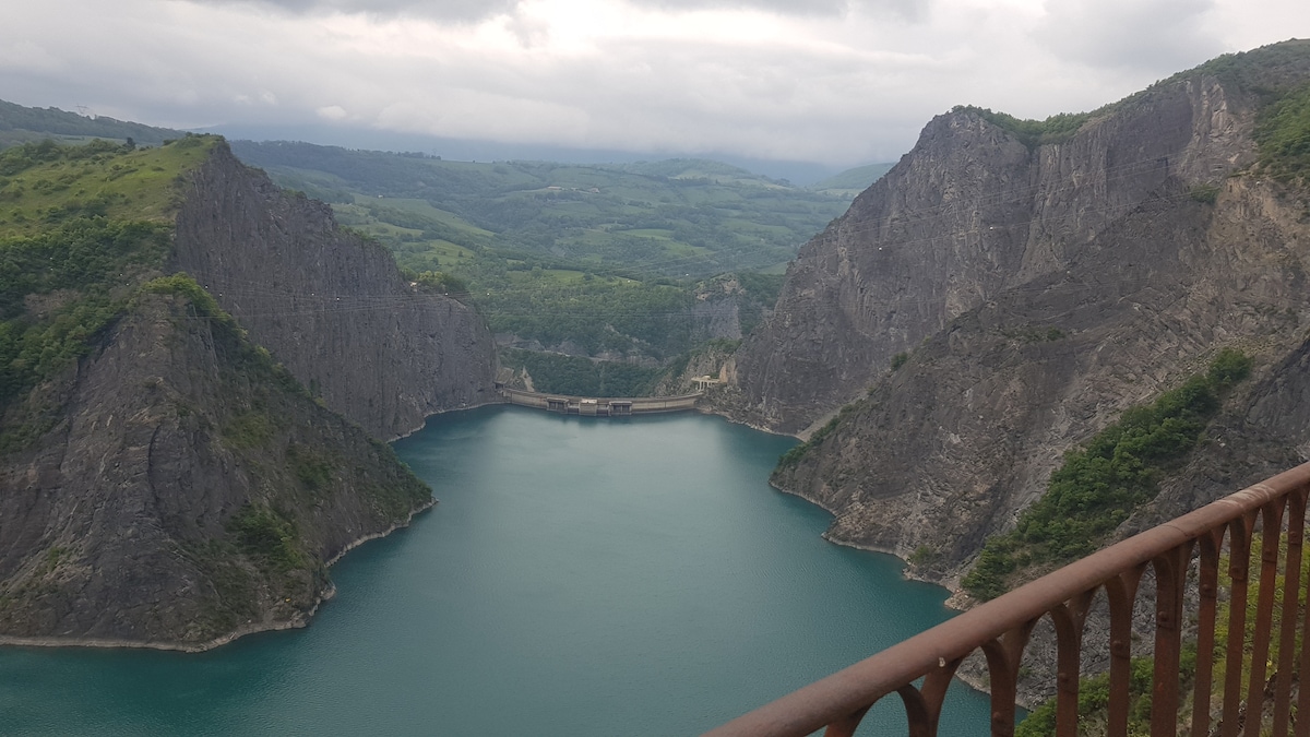 A scenic view of a tranquil blue lake surrounded by steep rocky cliffs and lush greenery. The water reflects the overcast sky, enhancing the serene atmosphere of the natural landscape. The image captures the impressive formations and detailed textures of the cliffs in the background.