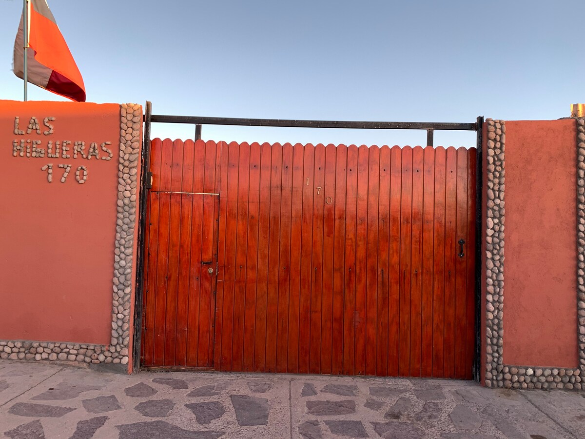 A vibrant red wooden gate marks the entrance to the property, framed by a stone wall. A smaller side door is integrated into the design, and a flag can be seen waving above. The textures and colors create a welcoming impression.