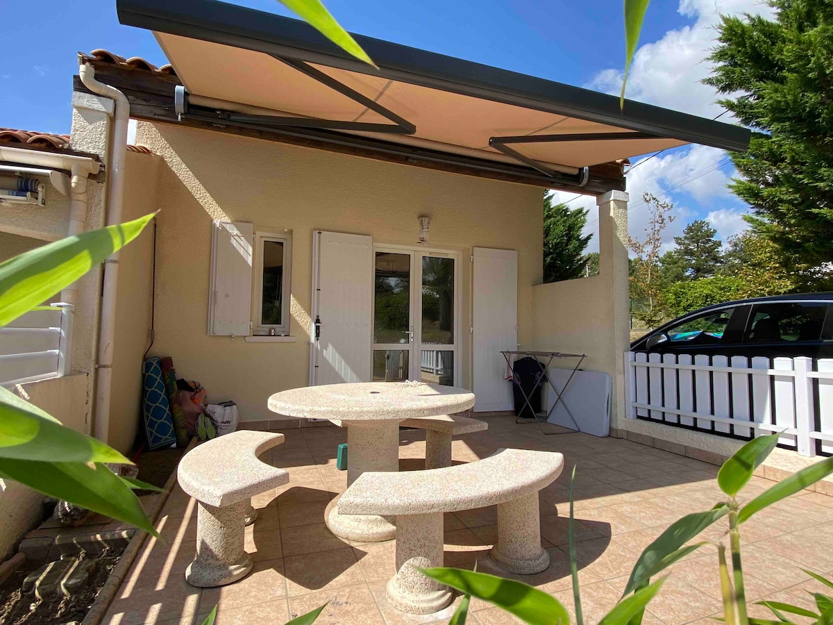 An outdoor terrace is shown, featuring a round stone table surrounded by curved seating. A retractable awning provides shade. The terrace is bordered by greenery and has a view of a white fence and a parked car.