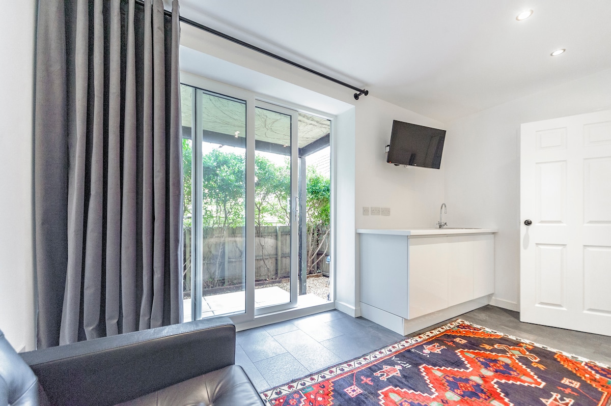 A sleek living area is highlighted by a modern kitchenette against a white wall. Large glass doors open to a small outdoor space, allowing natural light to fill the room. Dark curtains provide privacy, while a colorful area rug adds a touch of warmth to the flooring.