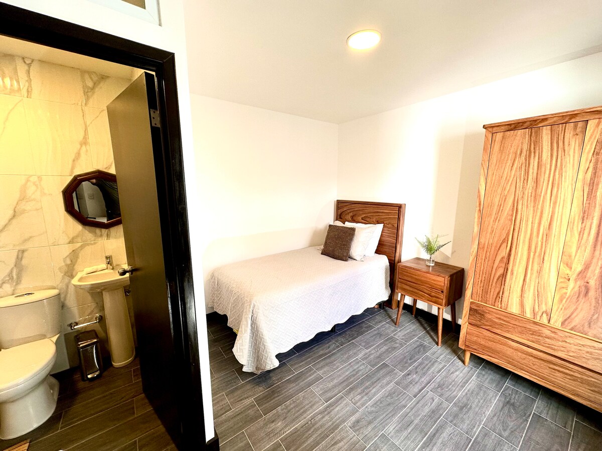 A compact bedroom features a simple bed with a light-colored coverlet. A wooden nightstand holds a decorative plant. The room includes a modern bathroom visible through an open door, showcasing minimalist fixtures and stylish tile work.