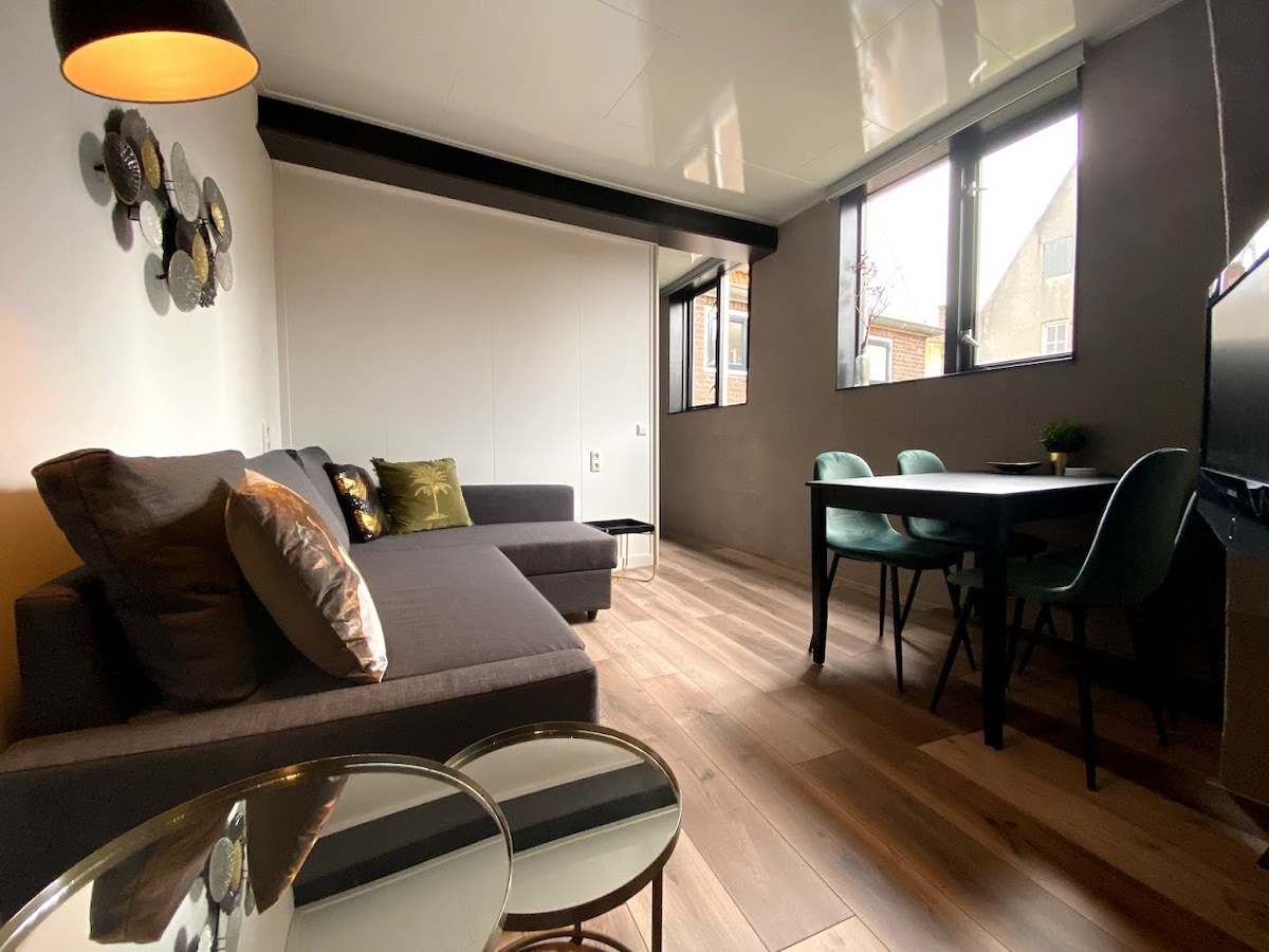 A modern living area is highlighted by a comfortable sectional sofa and a stylish dining table with green chairs. Large windows allow natural light to illuminate the space, while the polished wood floor adds warmth. Decorative elements are present on the wall.