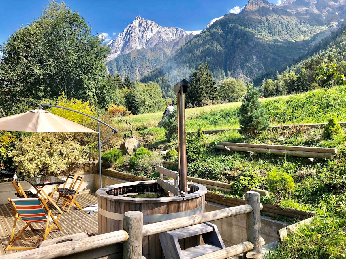 A wooden hot tub surrounded by a spacious deck offers scenic views of the mountains and greenery. Several lounge chairs are positioned nearby under a large umbrella, inviting relaxation in a serene outdoor space.
