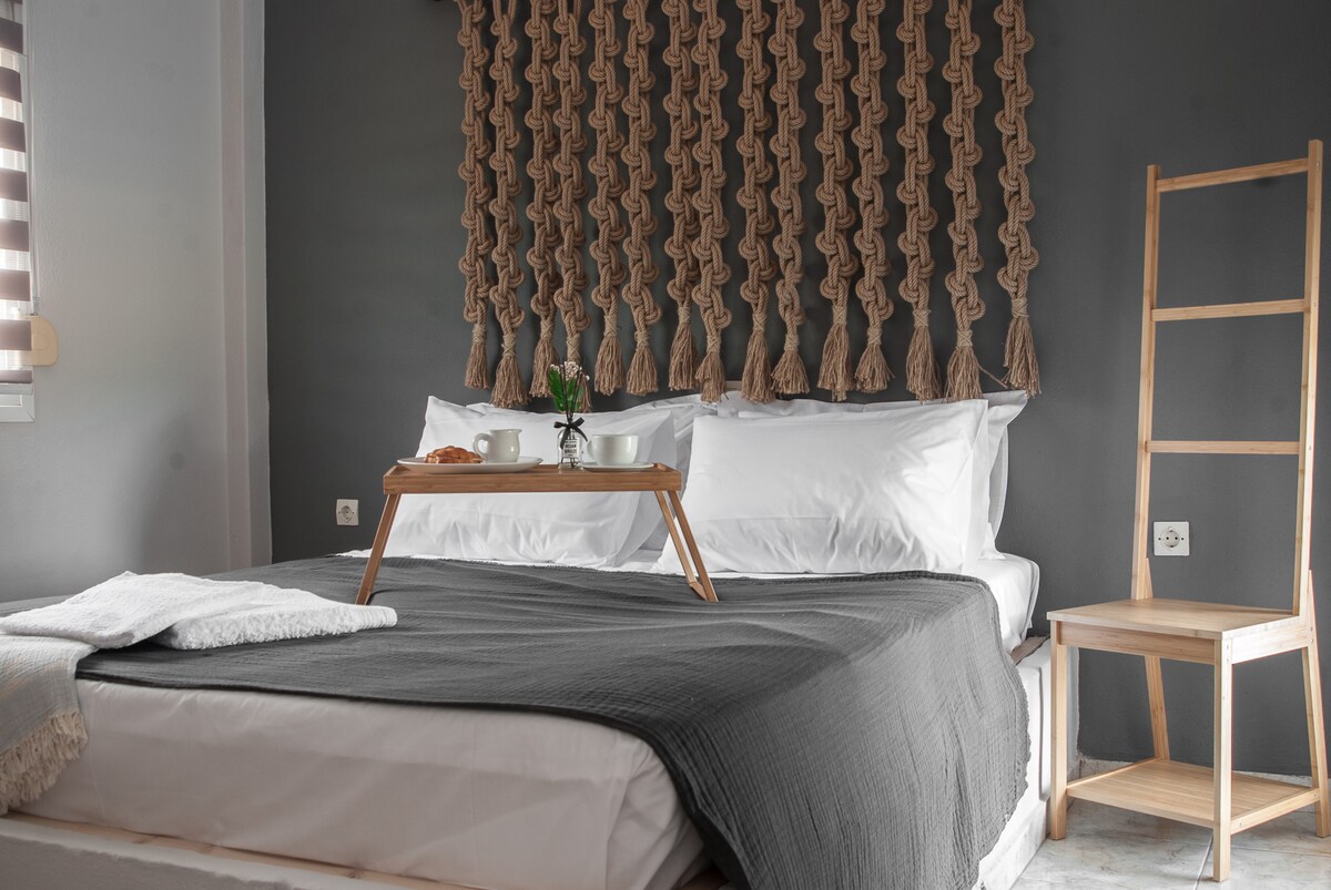 A spacious sleeping area features built-in beds adorned with a textured wall hanging made of natural fibers. A light blanket is draped across the bed, complemented by a portable tray on the bed holding a small plant and a pastry. A simple wooden chair is positioned nearby.