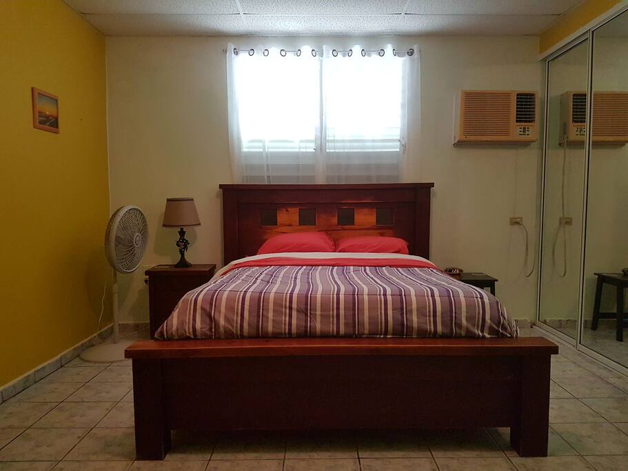 Master bedroom with queen size bed. The unit includes a private full bathroom and two walk-in closets. It has an A/C unit and a floor fan.
