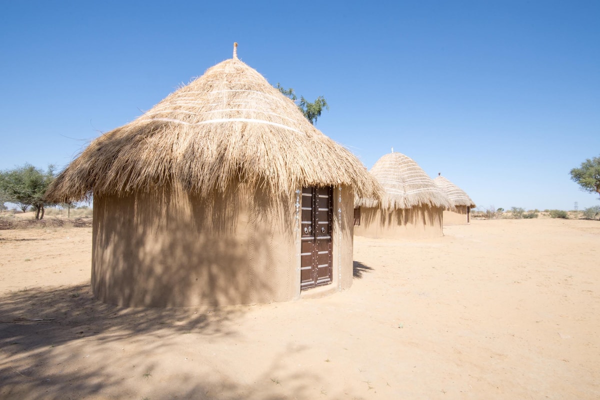 Traditional mud cottages with thatched roofs are positioned against a clear blue sky. The structures feature wooden doors and are surrounded by sandy terrain, providing a glimpse into the rustic desert living experience.