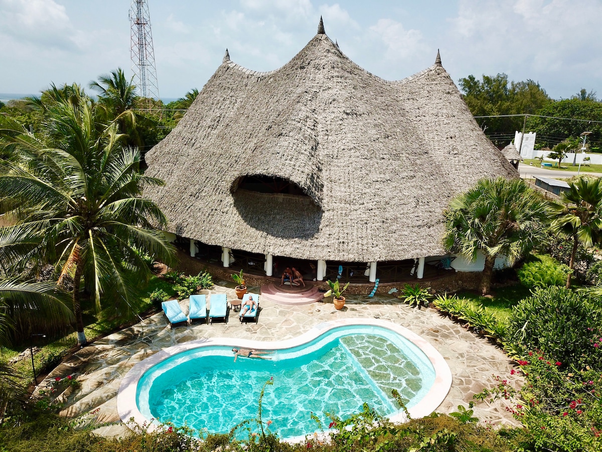The villa's unique Kenya-Style architecture features a thatched roof, blending harmoniously with its tropical surroundings. A private salt-water pool is framed by four sun-loungers and lush greenery, inviting relaxation and enjoyment under the sun.