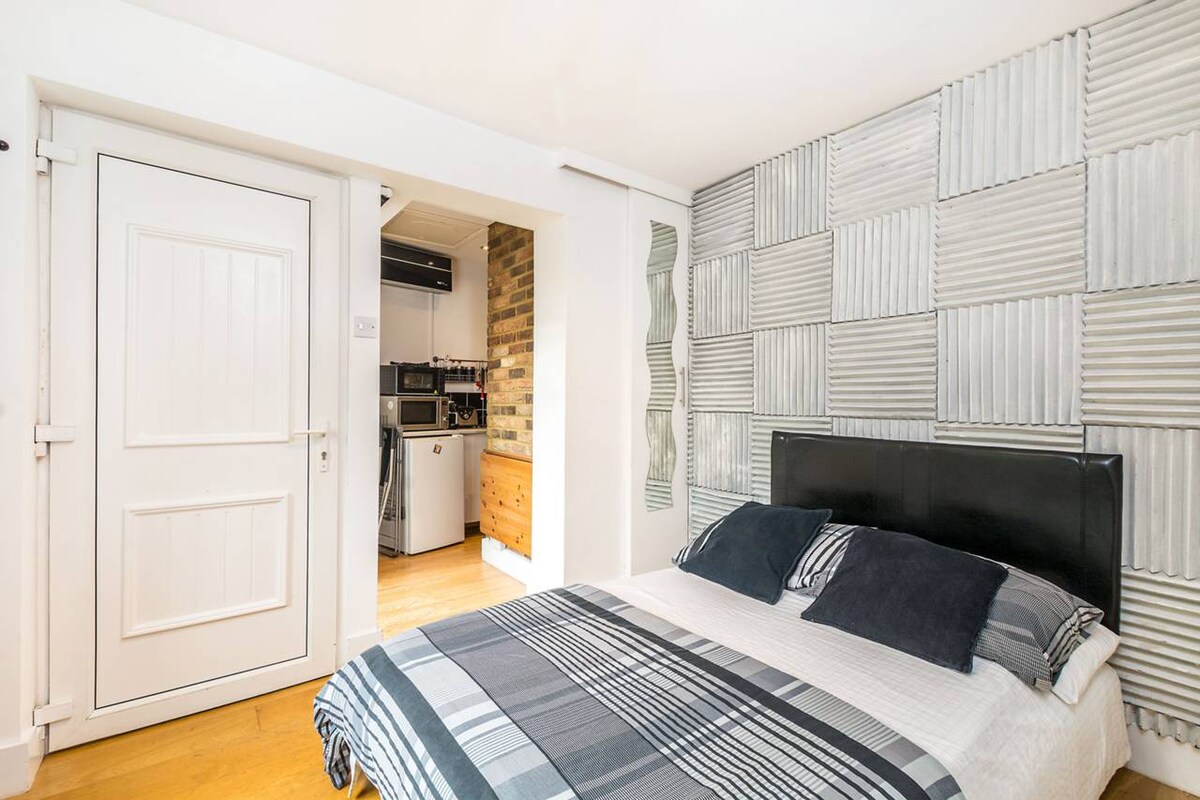 A studio room is presented with natural wood flooring and textured wall panels. A spacious bed with a dark headboard is dressed in a patterned blanket. A door leads to the kitchen area, which features modern appliances and exposed brick accents.