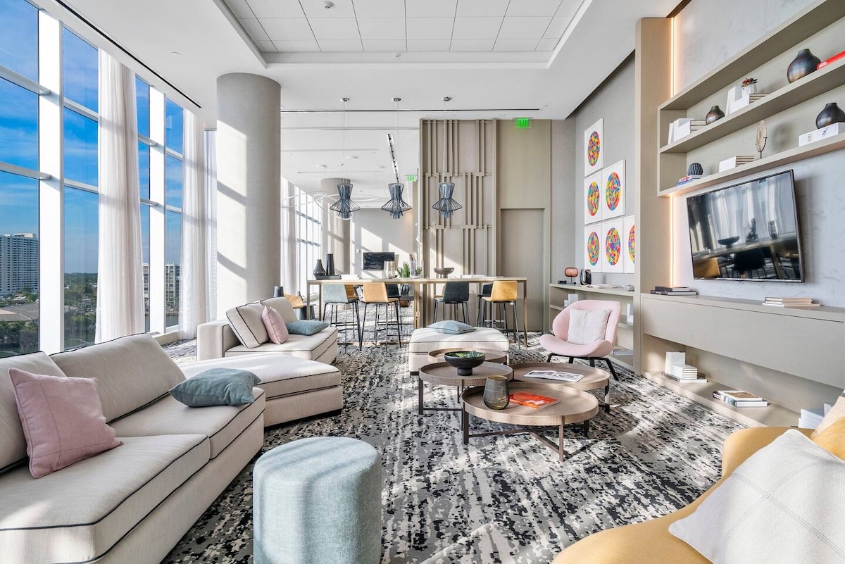 A modern lounge area is filled with natural light from large floor-to-ceiling windows. Soft seating in neutral tones complements colorful accent pillows, and a variety of tables are placed across a patterned rug. Artwork adorns the walls, adding visual interest to the space.