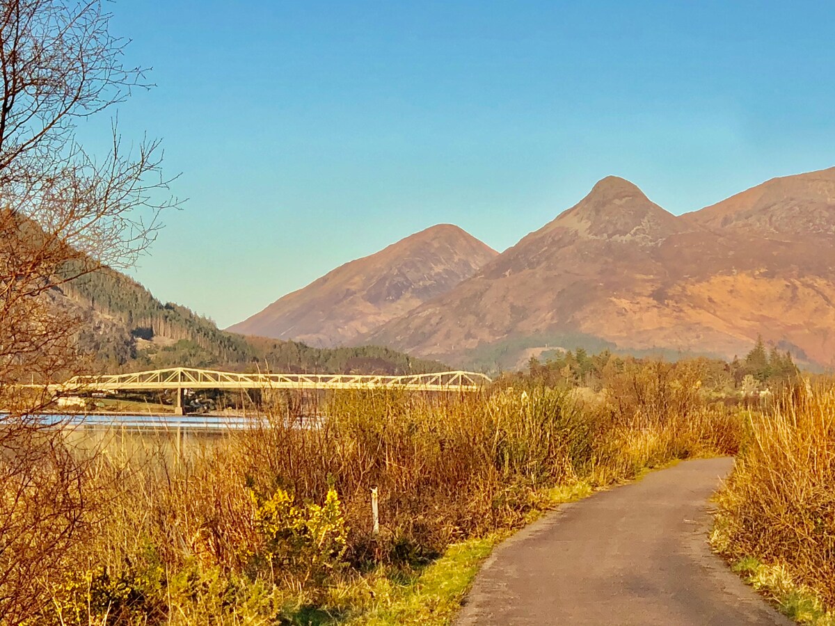 A scenic view reveals a winding path leading through tall grasses and shrubs, framed by distant mountainous terrain. A bridge spans across a body of water, under a clear blue sky, enhancing the natural beauty of the surrounding landscape.