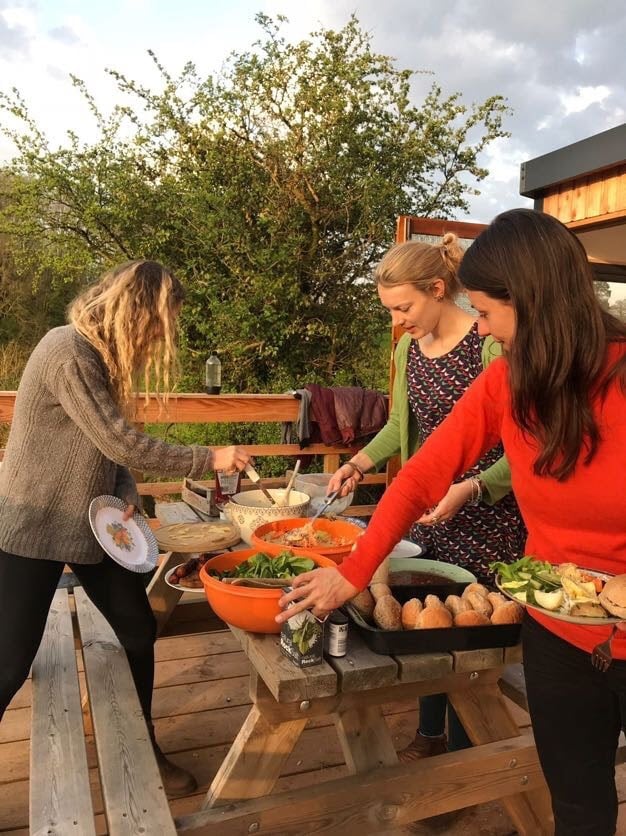 A communal dining setup is presented outdoors on a wooden deck, featuring a large table laden with colorful bowls of fresh salad and baked goods. Guests are engaged in food preparation, with lively greenery in the background enhancing the inviting atmosphere.