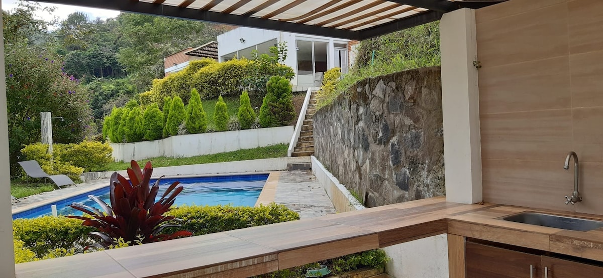 A well-appointed outdoor kitchen area overlooks a serene pool, framed by lush greenery and decorative shrubs. Natural stone elements and wooden surfaces are visible, adding to the overall aesthetic. A clear view of steps leading down to the pool area is also presented.