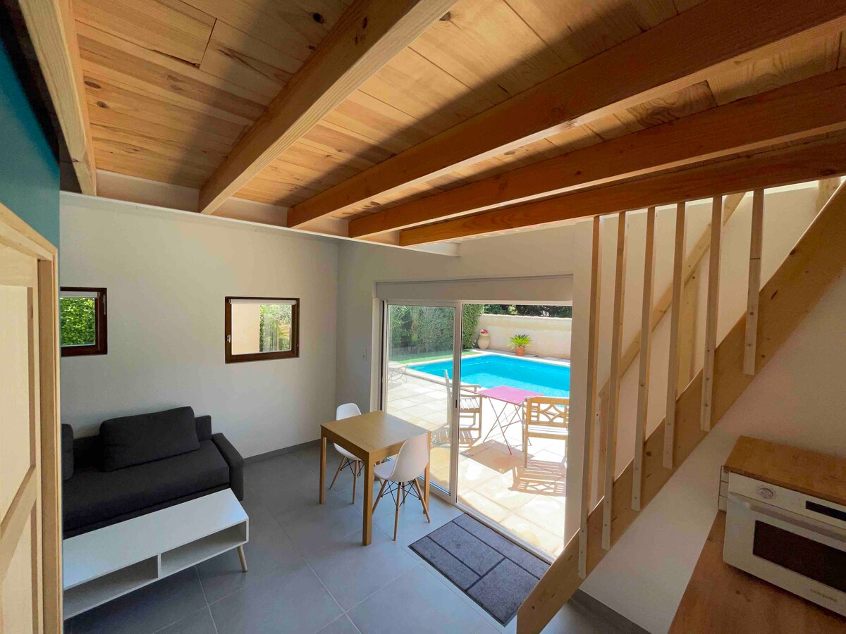 An open living area features a comfortable sofa, a small dining table, and sliding glass doors leading to a private terrace with pool access. Natural wood beams are visible on the ceiling, and light-colored walls enhance the spacious ambiance.