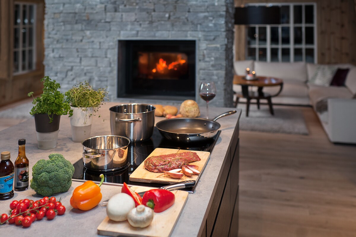 A spacious kitchen area is shown, featuring a large cooking island with various ingredients neatly arranged, including fresh vegetables and uncooked meat. Modern cookware, such as pots and a frying pan, rests on the induction cooktop. A cozy fireplace is visible in the background, providing warmth to the setting.