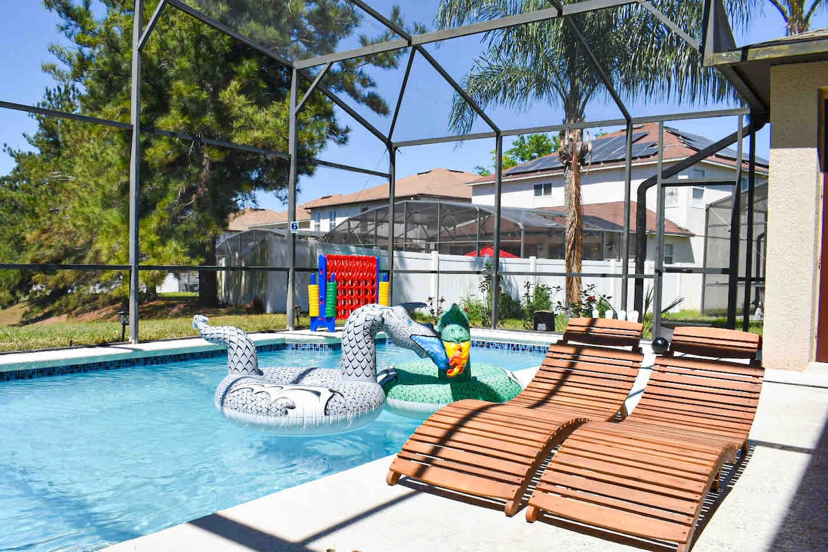 A refreshing pool area is enclosed by a mesh screen, featuring a blue swimming pool with colorful floatations. Two brown loungers are positioned nearby, inviting relaxation under the sun, while surrounding palm trees enhance the outdoor setting.