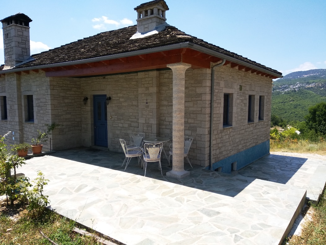 A stone exterior is complemented by a traditional tiled roof, showcasing a spacious patio area. Two metal chairs and a small table sit gracefully on the stone floor, with green hills visible in the background under a clear blue sky.