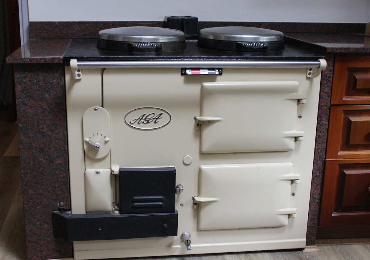 The heritage AGA stove is prominently displayed in a kitchen setting, featuring a cream-colored finish with rounded edges. Two metal cooking tops are visible, along with an adjacent wood cabinet, showcasing a blend of functional design and classic elegance.