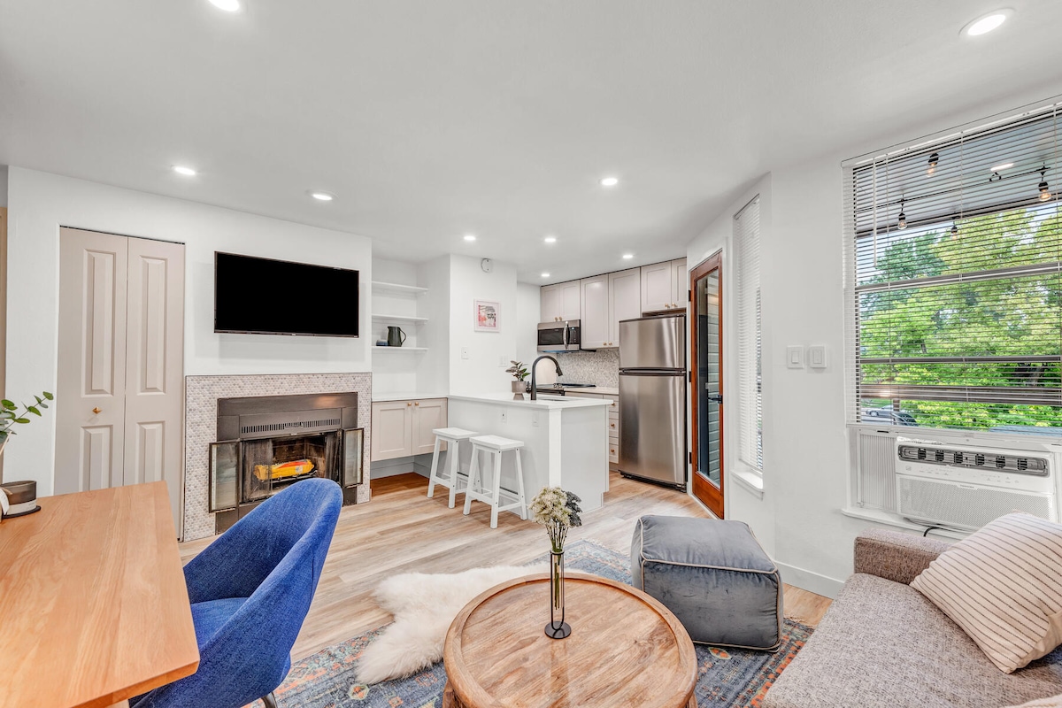 An open-concept living space features a modern kitchen with stainless steel appliances and a cozy sitting area. A fireplace adds warmth, while a large window provides natural light and views of greenery. Stylish furnishings include a round coffee table and a small desk.