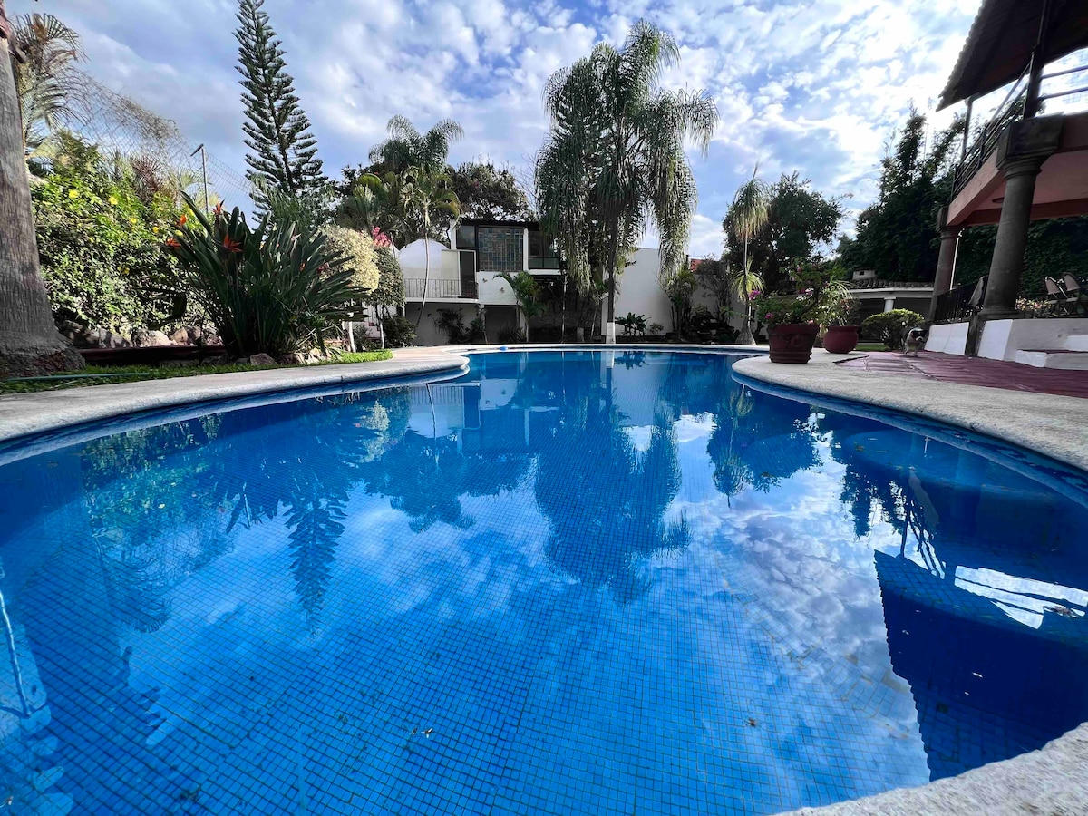 A crystal-clear pool is surrounded by lush greenery and colorful plants. The water reflects the sky, creating a serene atmosphere. The pool area is bordered by a smooth stone edge, inviting relaxation under a partly cloudy sky.