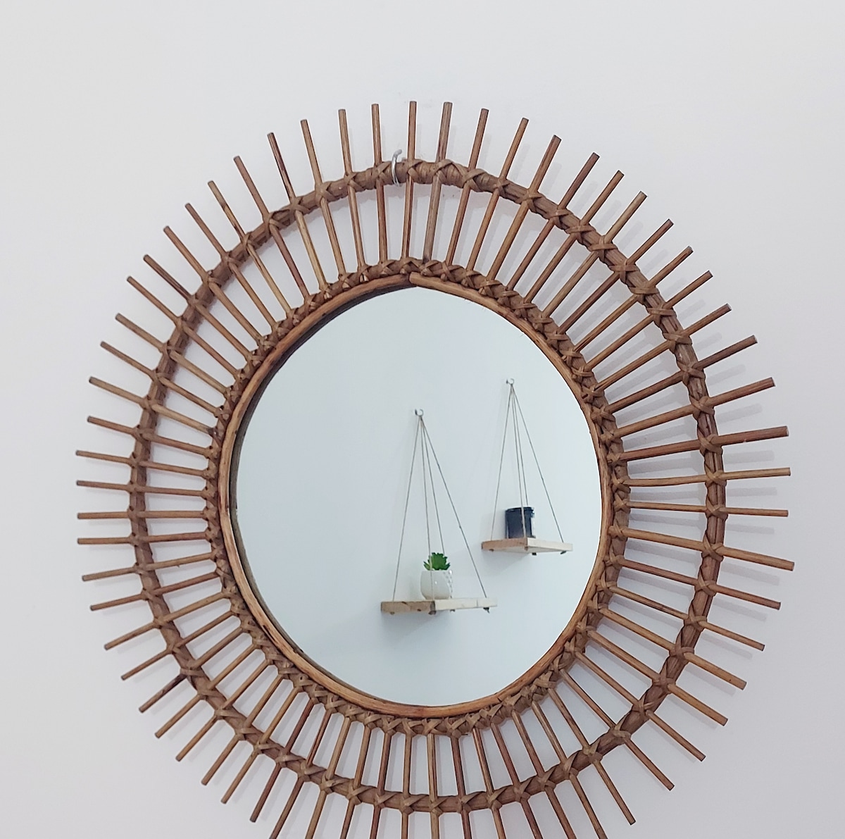 A circular mirror framed with a natural material showcases a sunburst design. The mirror reflects a minimalistic wall featuring two hanging shelves, one adorned with a small potted plant and a decorative object, creating an accent in the overall space.