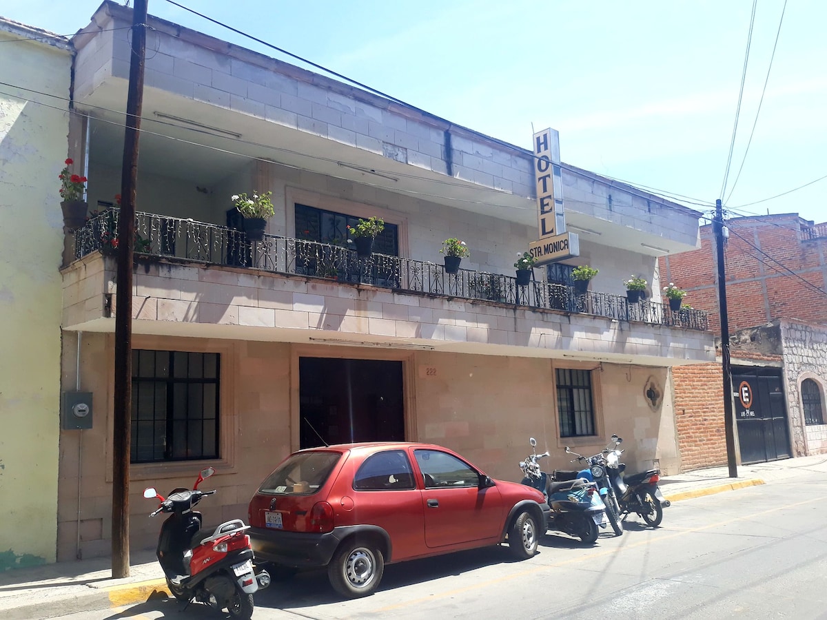 The hotel exterior features a two-story structure with a balcony adorned with potted plants. A red car and several motorcycles are parked in front, while the surrounding area shows a mix of urban architecture under a clear blue sky.