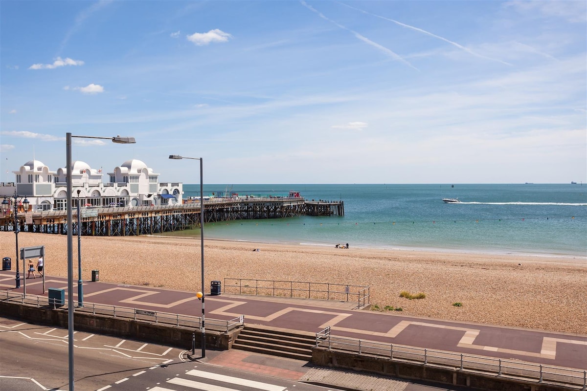 A clear view of the Southsea seafront features a sandy beach stretching before a wooden pier. Calm turquoise waters reflect the blue sky, while beachgoers enjoy the shoreline. The promenade is visible with street signs and lamp posts lining the street, adding to the coastal atmosphere.