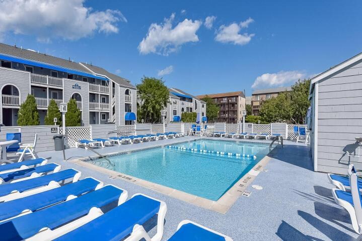 A well-maintained swimming pool is surrounded by blue lounge chairs. The pool area is enhanced by a pristine water surface, with several umbrellas providing shade. Nearby buildings with balconies are visible against a clear sky dotted with clouds.