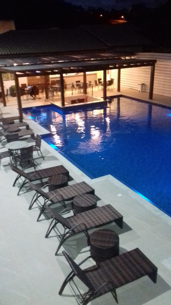 An extensive swimming pool is illuminated with soft lighting, reflecting a calm blue hue. Numerous lounge chairs surround the pool, arranged with small side tables. A covered area in the background suggests dining options, visible through large windows.