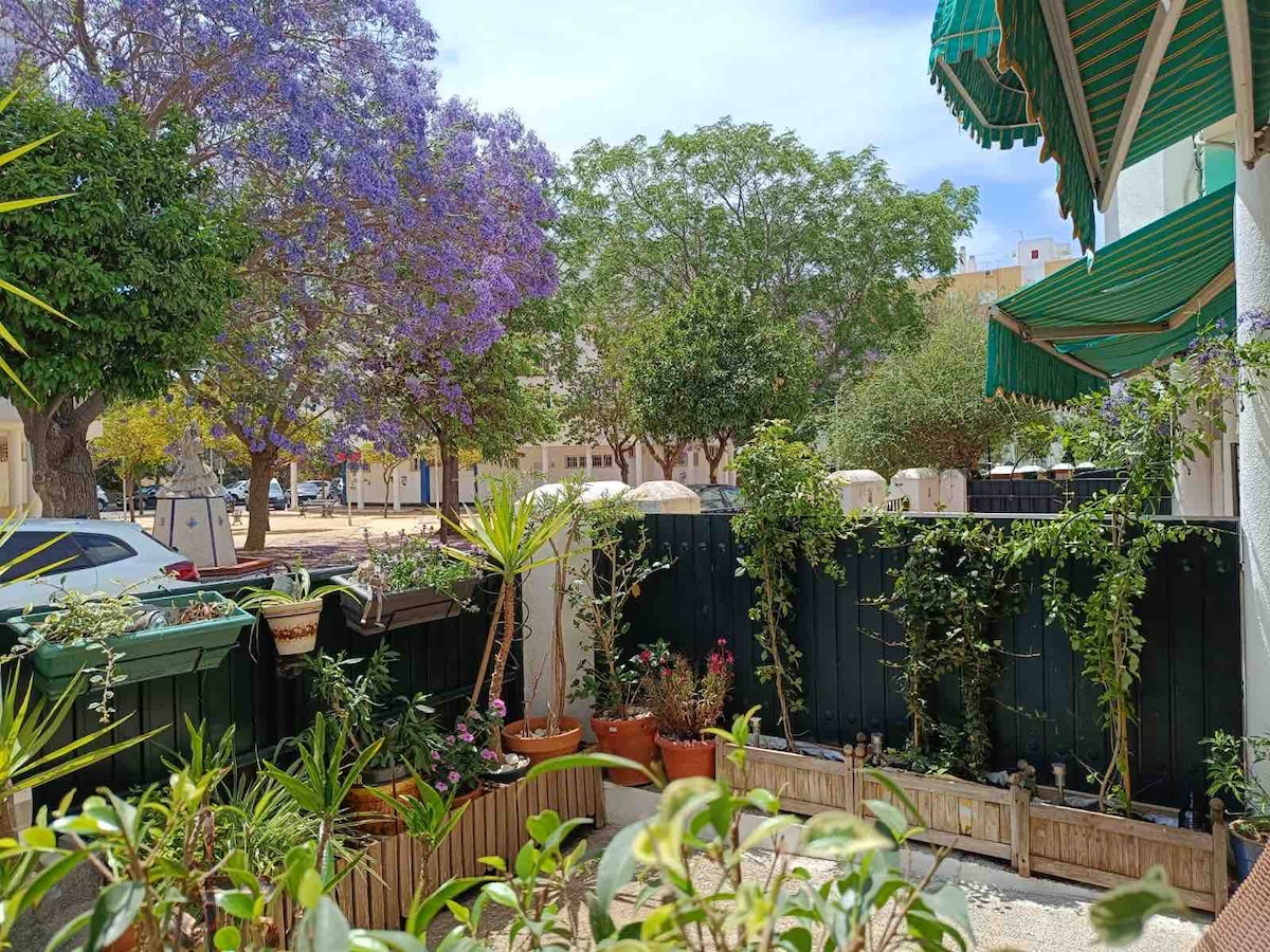 A lush garden area is displayed, featuring various potted plants and flowering shrubs. The greenery is complemented by the vibrant purple blossoms of nearby trees. A light green awning can be seen overhead, providing shade to the outdoor space.