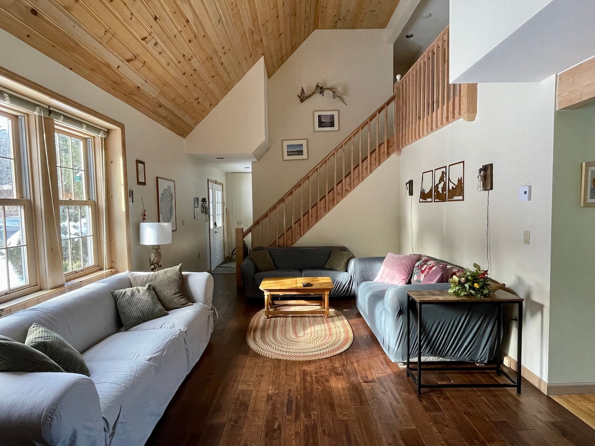 A well-lit living area features a high wood-paneled ceiling and a mix of seating options, including a light-colored sofa and a darker sectional. A wooden coffee table sits atop a circular rug, and decorative artwork is arranged along the walls.