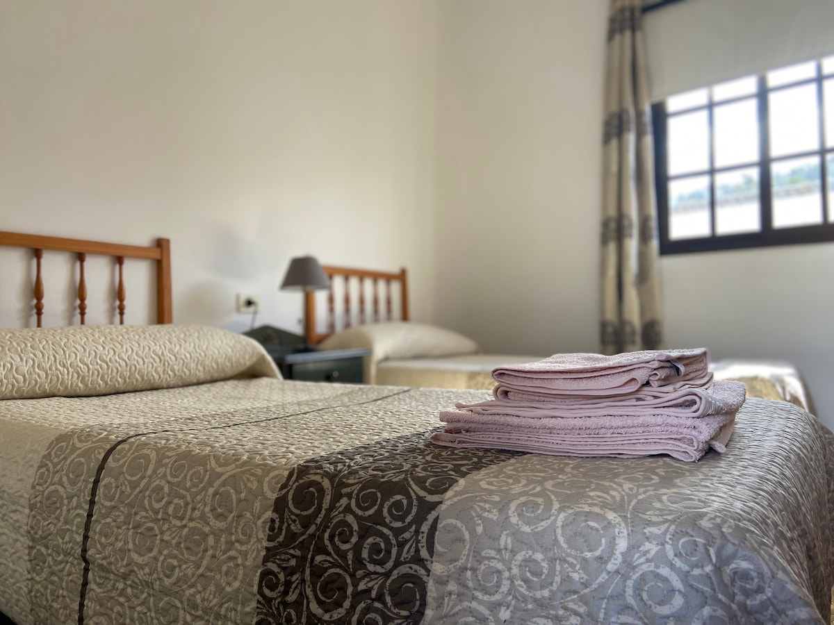 A serene bedroom setting is presented with two beds featuring textured bedding. A neatly arranged stack of towels is placed on one bed. Soft natural light filters through a window, which is framed by light curtains, enhancing the calming environment.