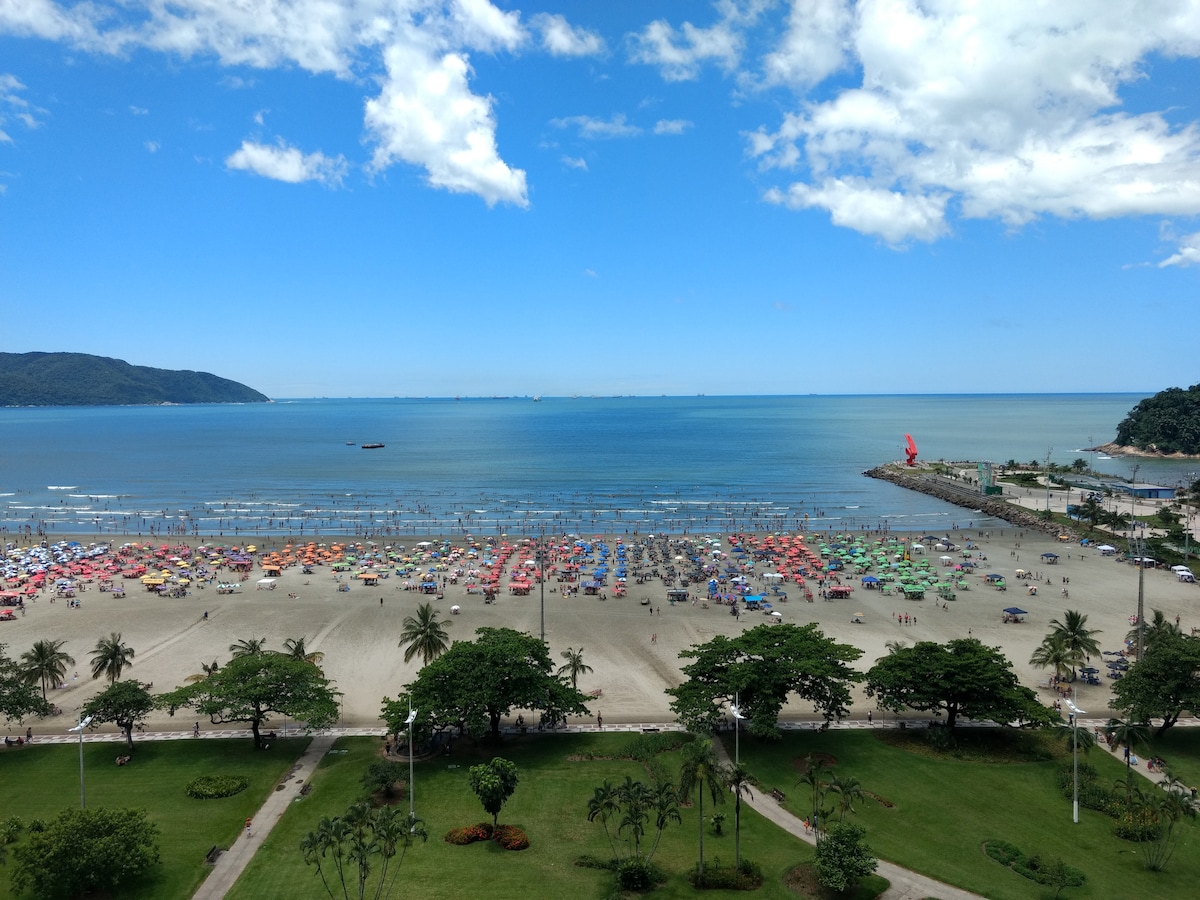 A panoramic view of the beach is presented, featuring numerous colorful umbrellas on the sandy shore. The blue waters meet a clear sky dotted with a few clouds. Lush green areas with palm trees and colorful patches of vegetation enhance the natural scenery.