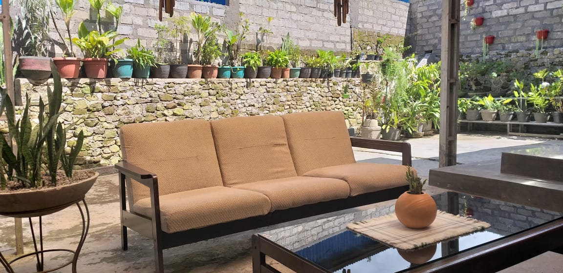 A shaded outdoor seating area features a brown fabric sofa positioned in front of a stone wall adorned with potted plants. A low glass table rests on the patio, complementing the natural surroundings with decorative elements.