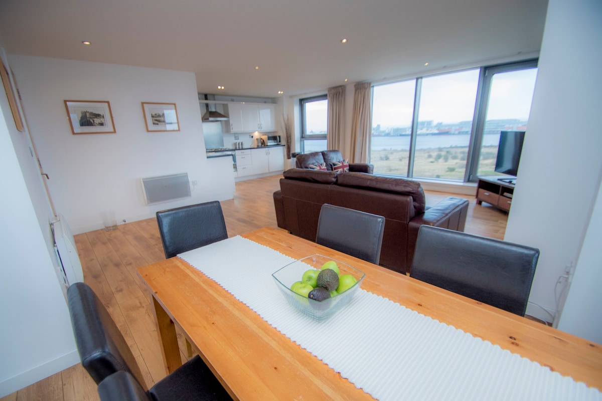 An inviting open living area features a wooden dining table set for six, adorned with a centerpiece of fresh fruit. The spacious lounge displays comfortable seating and large windows that offer expansive views of the surrounding landscape.