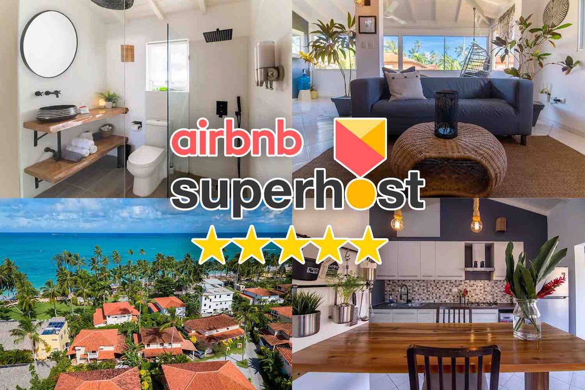 An airy bathroom is highlighted by modern fixtures and a circular mirror. A cozy living space features a gray sofa and a stylish coffee table. An aerial view reveals the surrounding beachside area. A sleek kitchen boasts wood accents and pendant lighting, emphasizing contemporary design.
